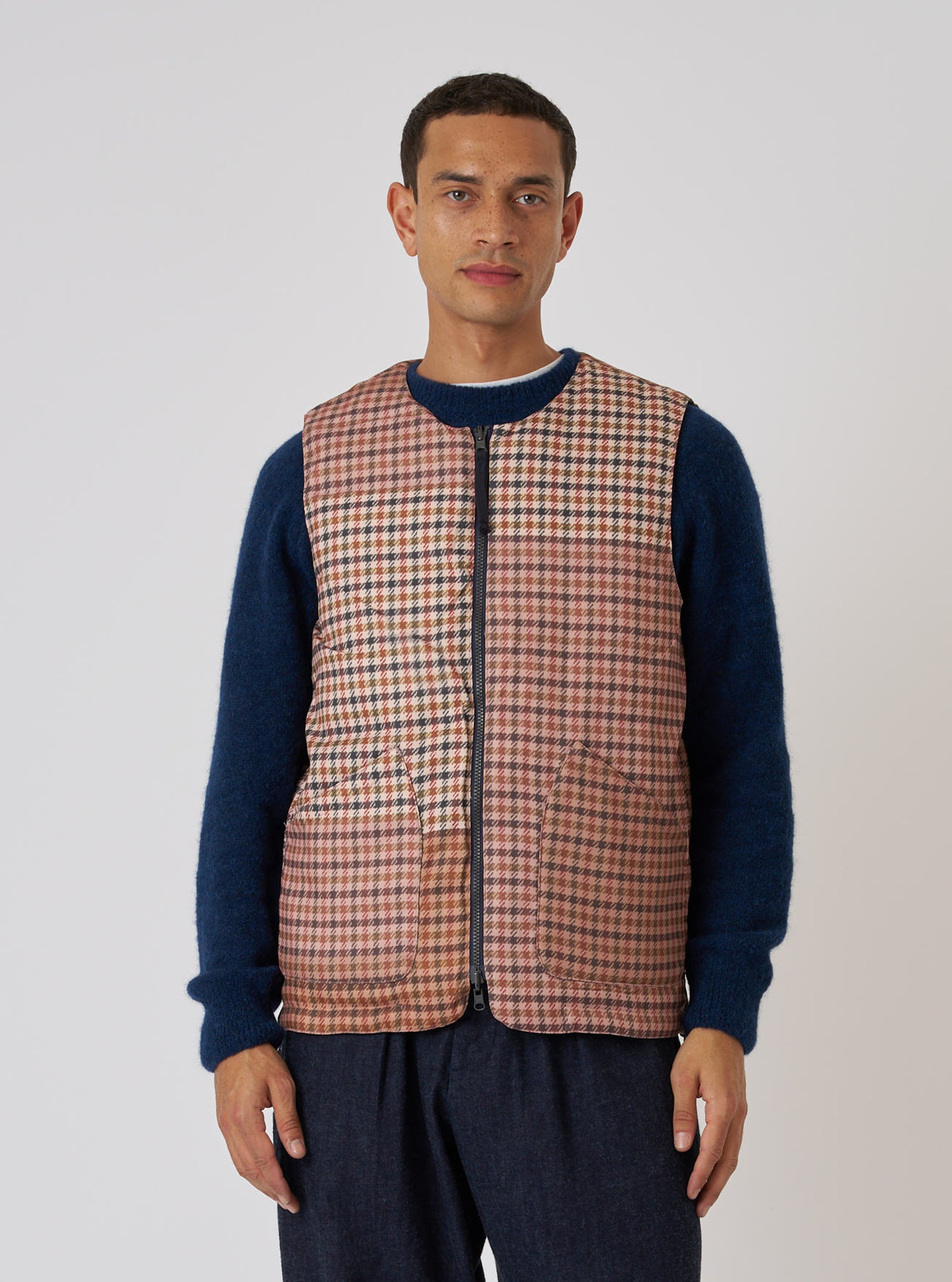 A model poses in front of a white background, wearing the TAION x Universal Works Reversible Dark Navy Military Line Vest. The vest is brown and tan plaid with a zip front.