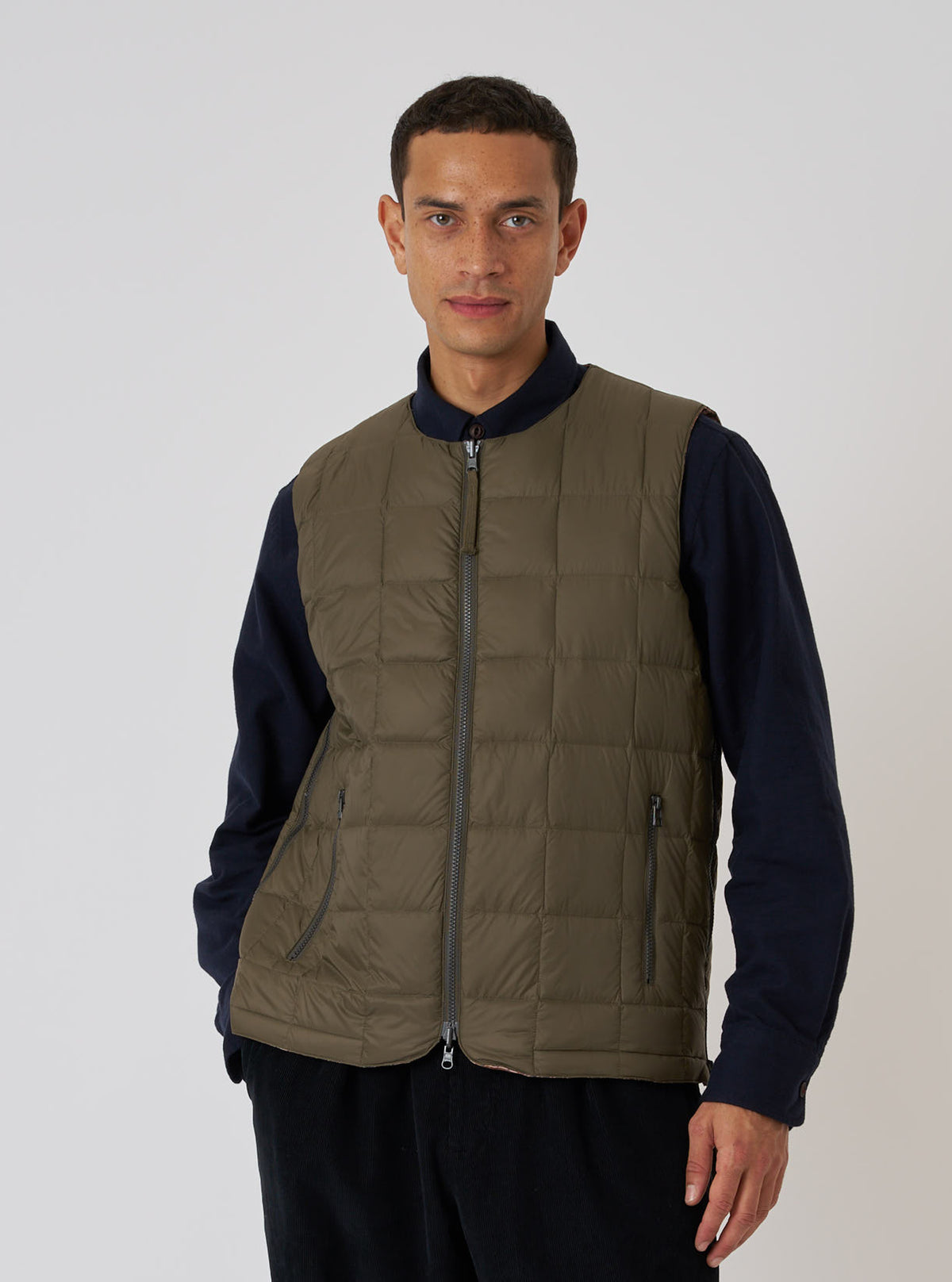 A model wears the TAION x Universal Works Reversible Dark Olive Military Line Vest. The quilted vest is dark olive with a full zip. Shot in a studio with a white background.