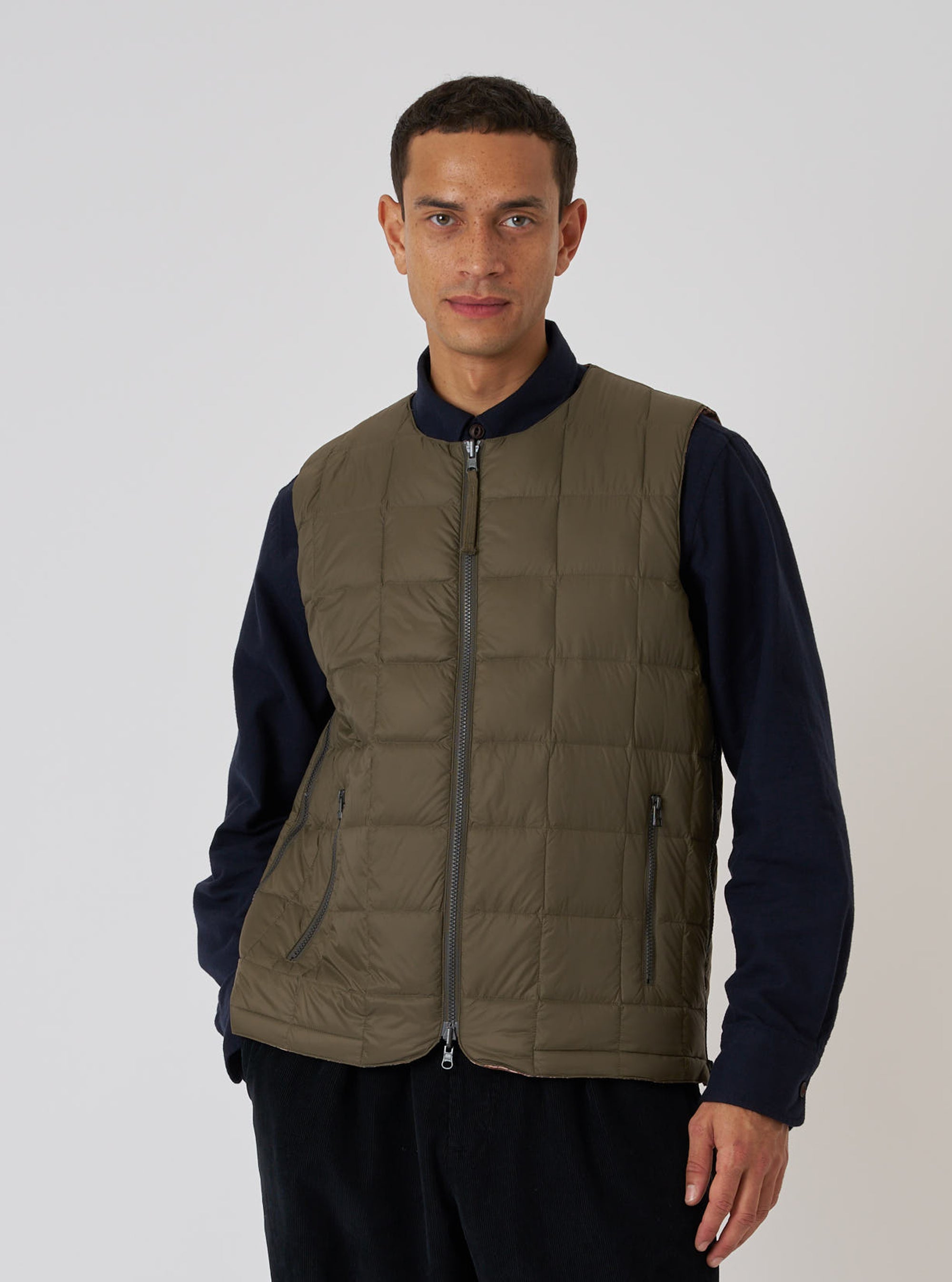 A model wears the TAION x Universal Works Reversible Dark Olive Military Line Vest. The quilted vest is dark olive with a full zip. Shot in a studio with a white background.