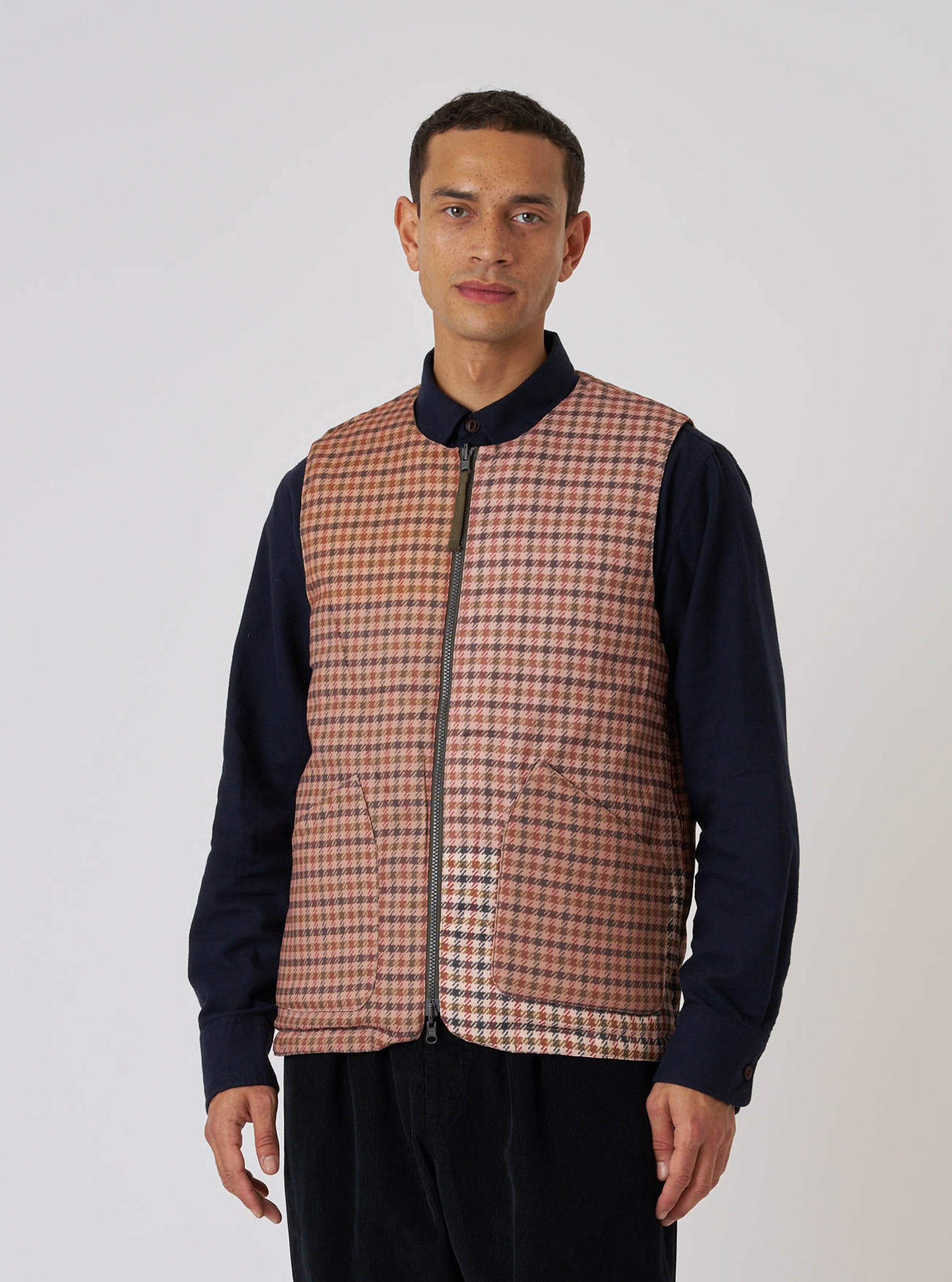 A model on a white background wears the TAION x Universal Works Reversible Dark Olive Military Line Vest. The vest is a brown and tan houndstooth pattern with a zip front.
