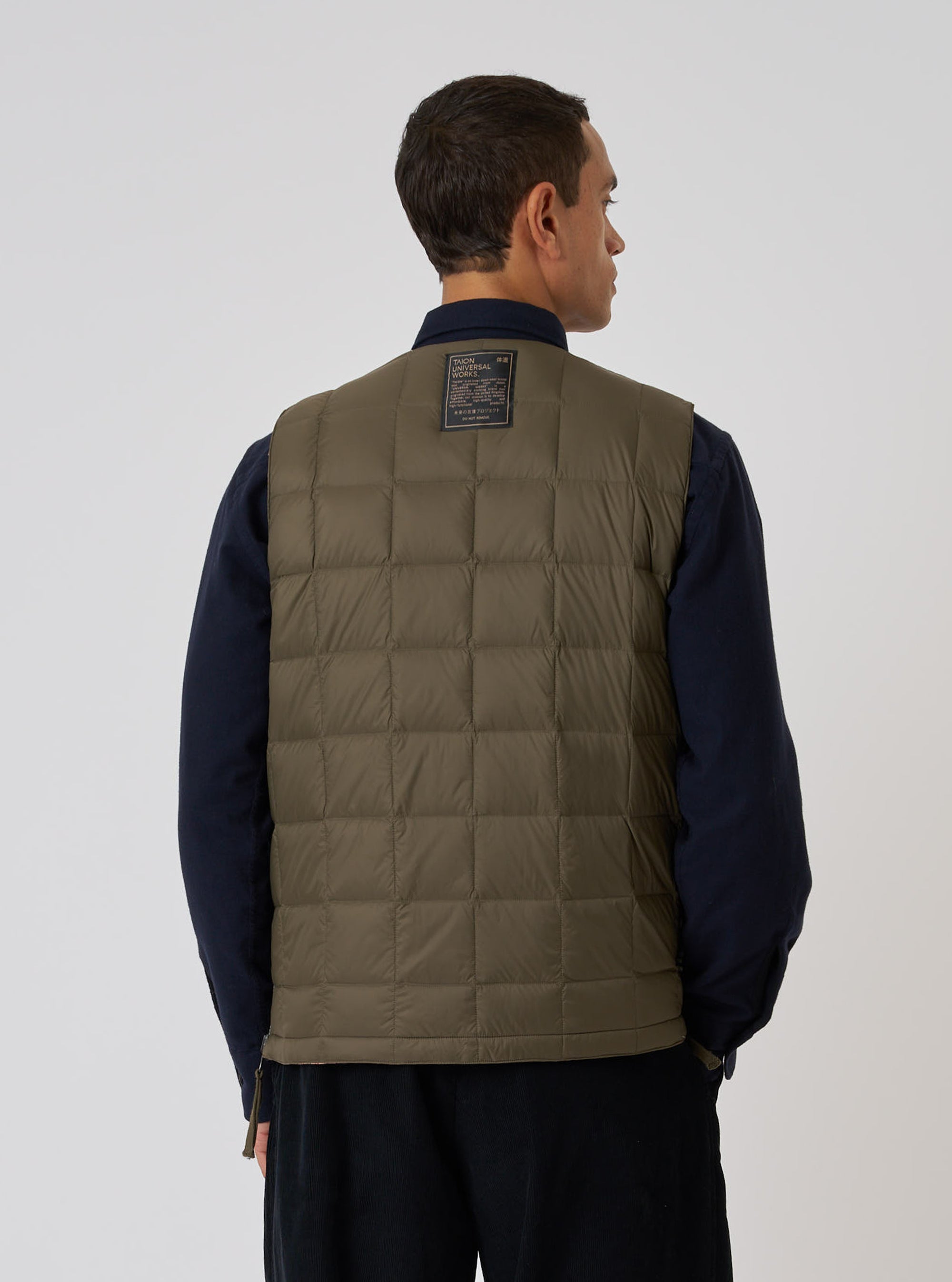 Back view of a model wearing a TAION x Universal Works Reversible Dark Olive Military Line Vest. The quilted vest is styled over a navy shirt, against a plain white backdrop.