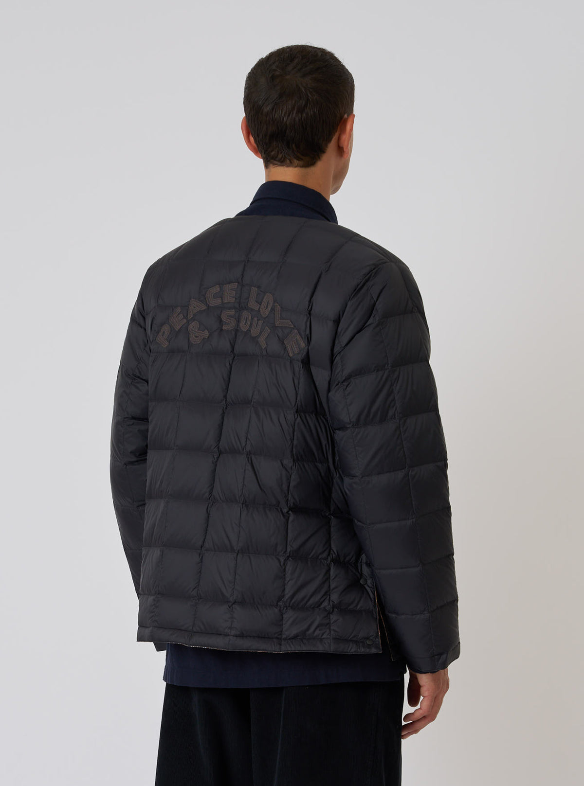 Back view of a model wearing the TAION x Universal Works Reversible Black Military Line Liner Jacket. The quilted jacket has 'Peace Love & Soul' embroidered on the back. White background.