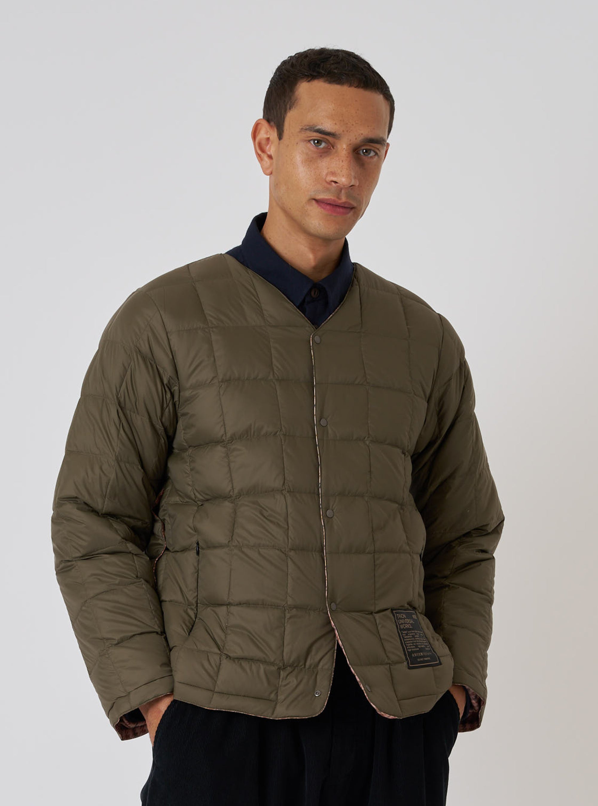 A model in a TAION x Universal Works Reversible Dark Olive Military Line Liner Jacket. The quilted jacket is on a white background. The fit is relaxed.