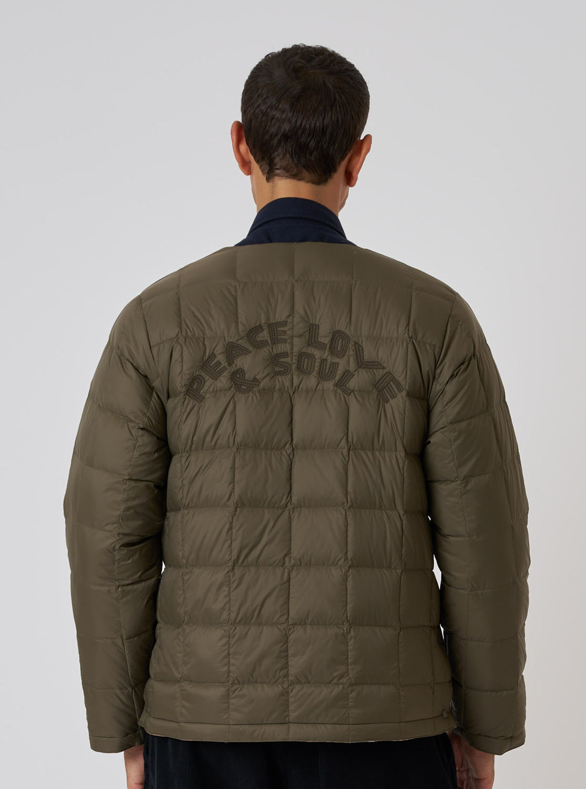 Back view of a model wearing the TAION x Universal Works Reversible Dark Olive Military Line Liner Jacket against a white background. The jacket is quilted with 'PEACE LOVE & SOUL' on the back.