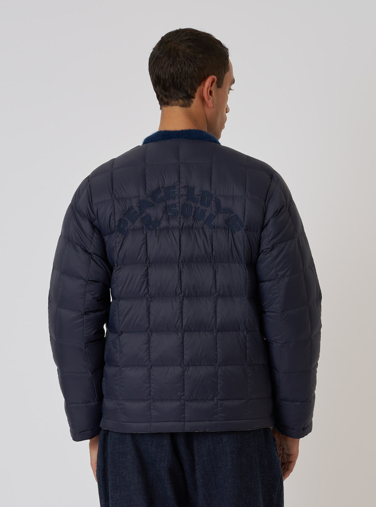 Back view of a model wearing the TAION x Universal Works Reversible Dark Navy Military Line Liner Jacket. The quilted jacket has 'PEACE LOVE & SOUL' embroidered on the back. Clean white background.