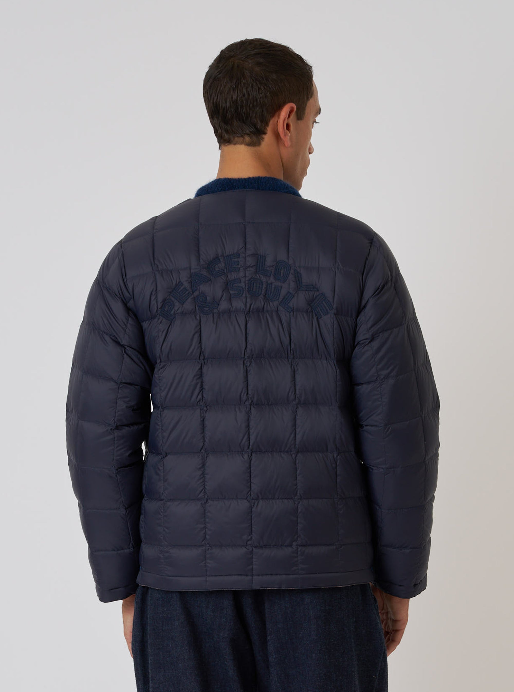 Back view of a model wearing the TAION x Universal Works Reversible Dark Navy Military Line Liner Jacket. The quilted jacket has 'PEACE LOVE & SOUL' embroidered on the back. Clean white background.