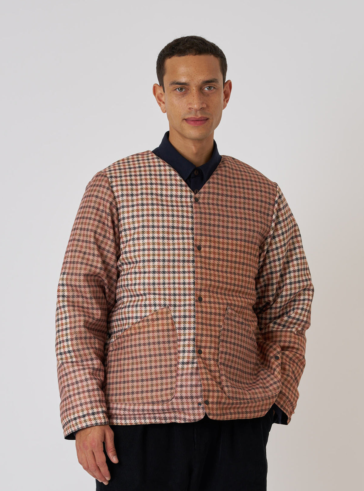 A model poses in front of a white background wearing the TAION x Universal Works Reversible Dark Olive Military Line Liner Jacket. The jacket is a brown and tan houndstooth pattern.