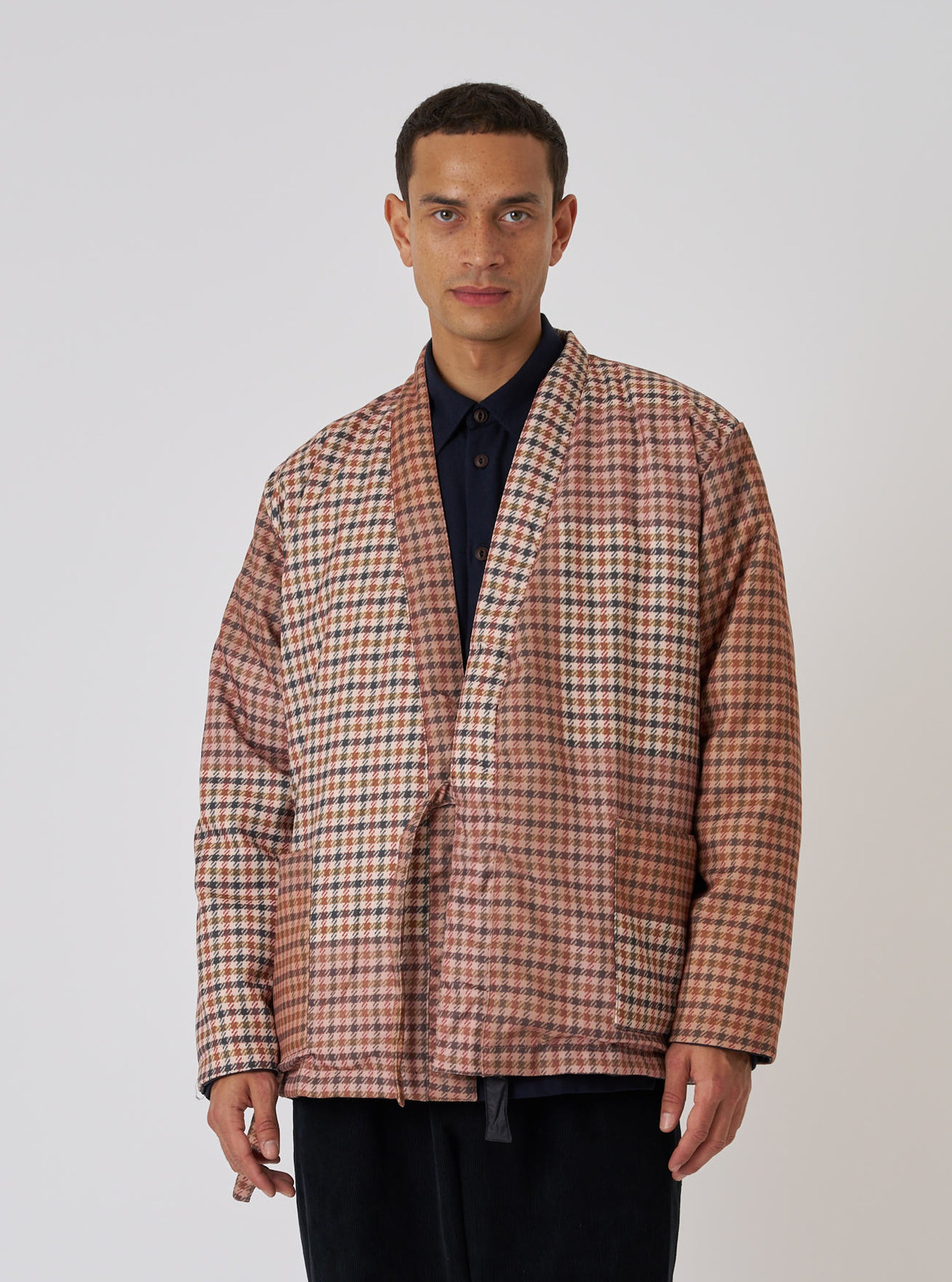 A model wears the TAION x Universal Works Reversible Dark Olive Military Line Kyoto Work Jacket. The jacket has a brown, tan, and green check pattern. Shot on a white background.