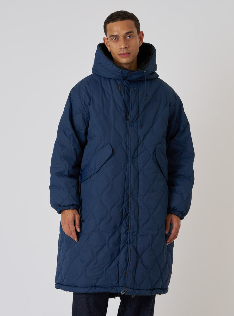 A model poses in front of a white background wearing the TAION x Universal Works Reversible Dark Navy Military Line Sherpa Parka. The parka is quilted and has a sherpa-lined hood.