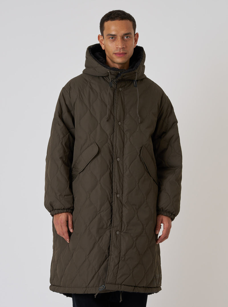 A model poses in front of a white background, wearing a TAION x Universal Works Reversible Dark Olive Military Line Sherpa Parka. The parka is quilted and has a sherpa lining.