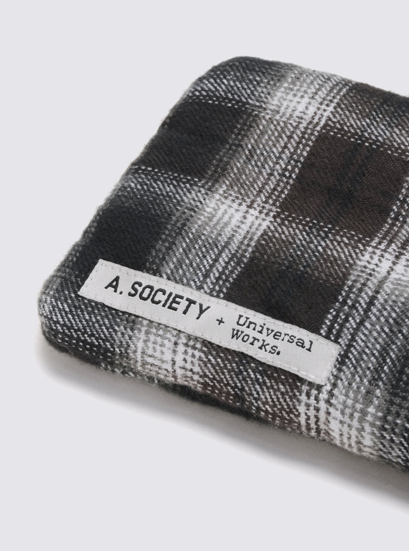 Close-up of A. SOCIETY x Universal Works Olive/Green Kowloon Eyewear case. Plaid pattern in olive and green hues.  A white label with the brand names is stitched on. Shot on a white surface.