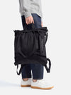 A person holds the F/CE.® Black Recycled Twill 3-Way Helmet Bag in a studio shot. The bag is black, made of twill, and has straps. The person wears jeans and white sneakers.