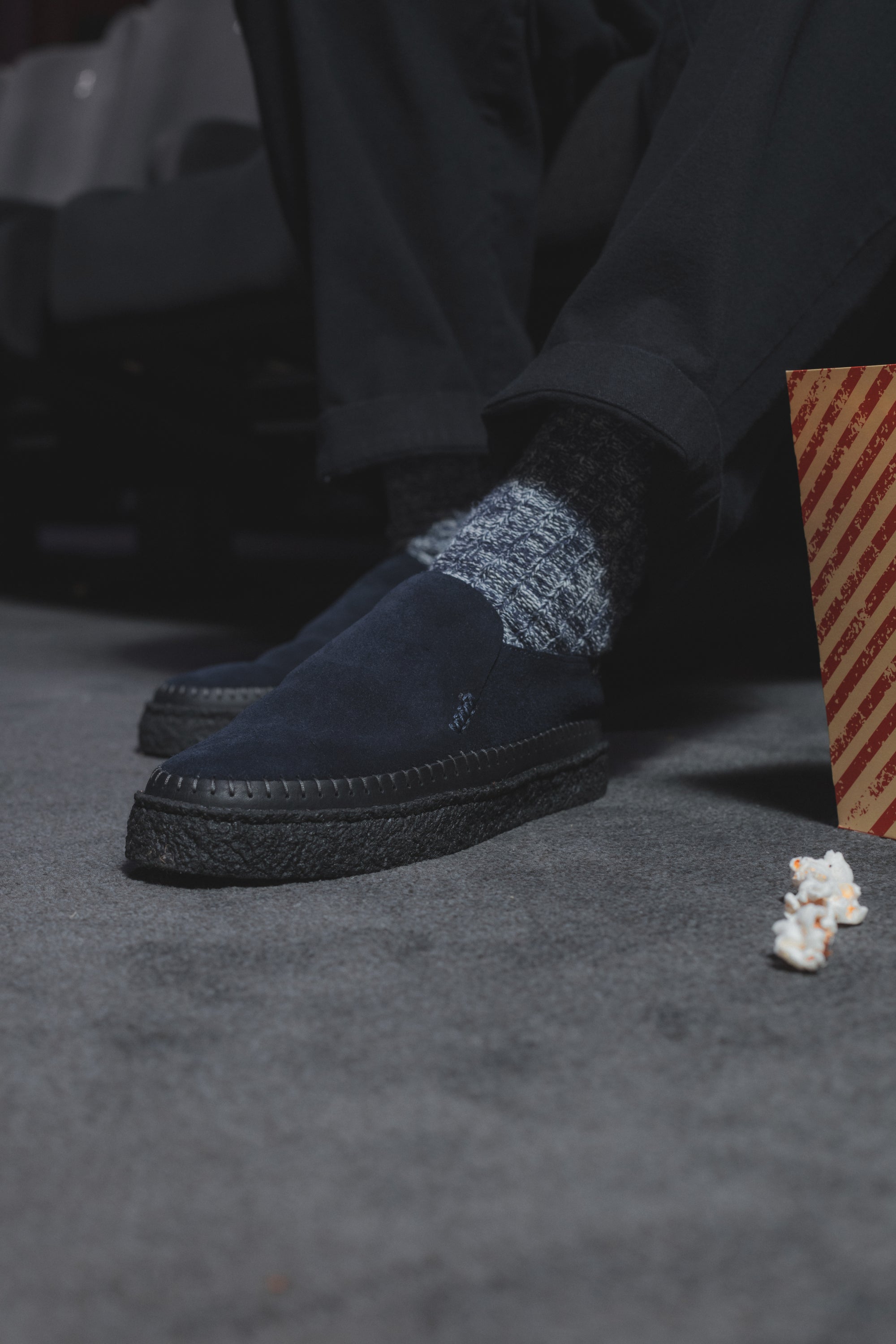 Person wearing Yogi x Universal Works Hitch Loafer in Indigo Suede/Crepe, with grey socks. The shoes are indigo suede with a black crepe sole. Popcorn and a popcorn box are on the grey carpet.