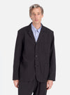 A model poses in a Charcoal Fine Linen Mix Three Button Jacket against a white backdrop. The jacket is paired with a light blue shirt. The fit is relaxed and modern.