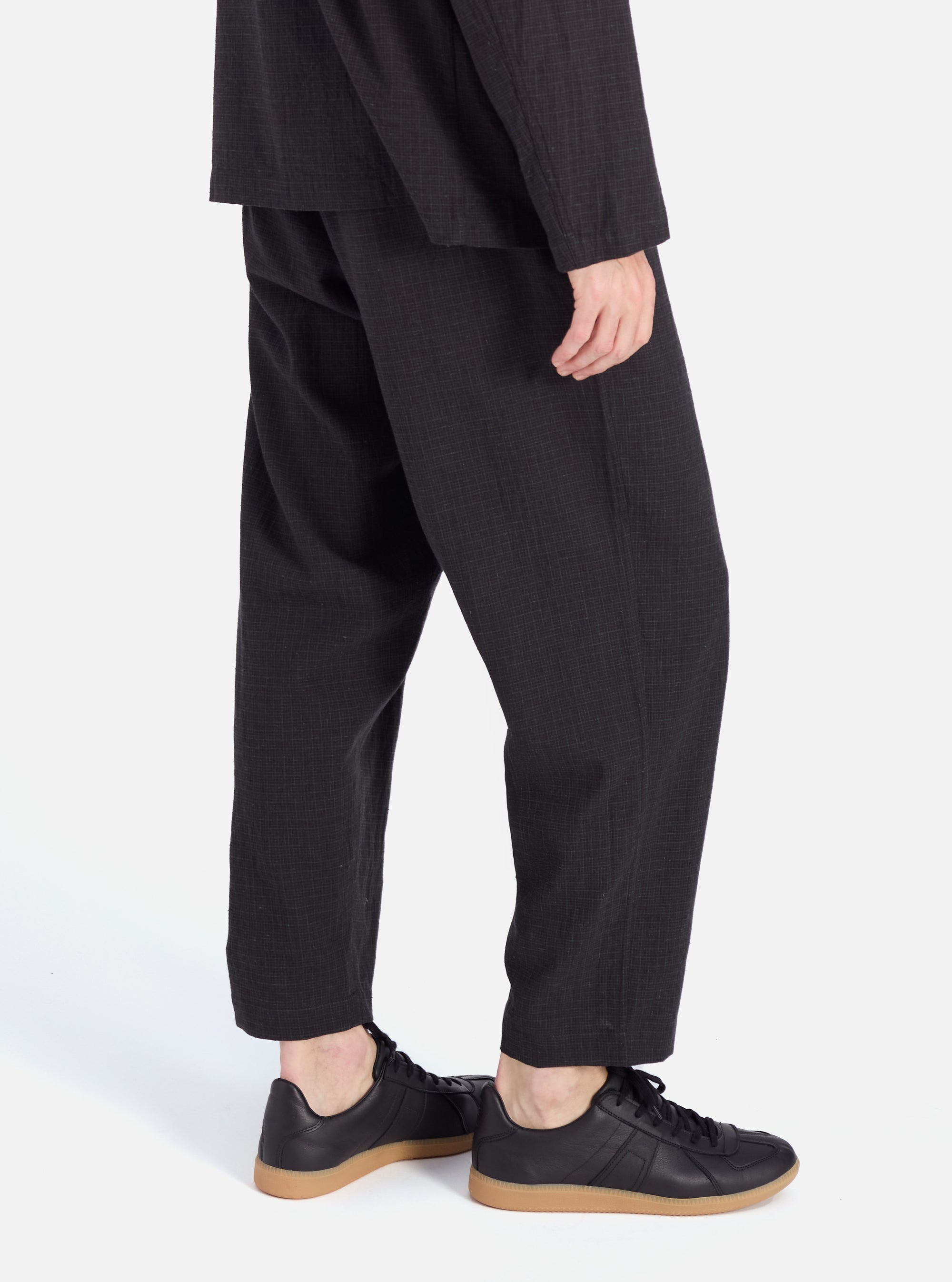 Side view of a model wearing Charcoal Fine Linen Mix Pleated Track Pant. The pants are paired with black sneakers. The background is a clean, bright white.