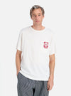 A model poses in an Ecru/Red Single Jersey Print Pocket Tee against a white background. The tee is paired with striped pants. The pocket has a red print.