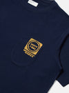 Close-up of the Navy/Yellow Single Jersey Print Pocket Tee on a white surface. The navy tee features a yellow print on the pocket. 