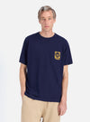 A model poses in a Navy/Yellow Single Jersey Print Pocket Tee against a white background. The tee is paired with khaki pants. The pocket has a yellow print.