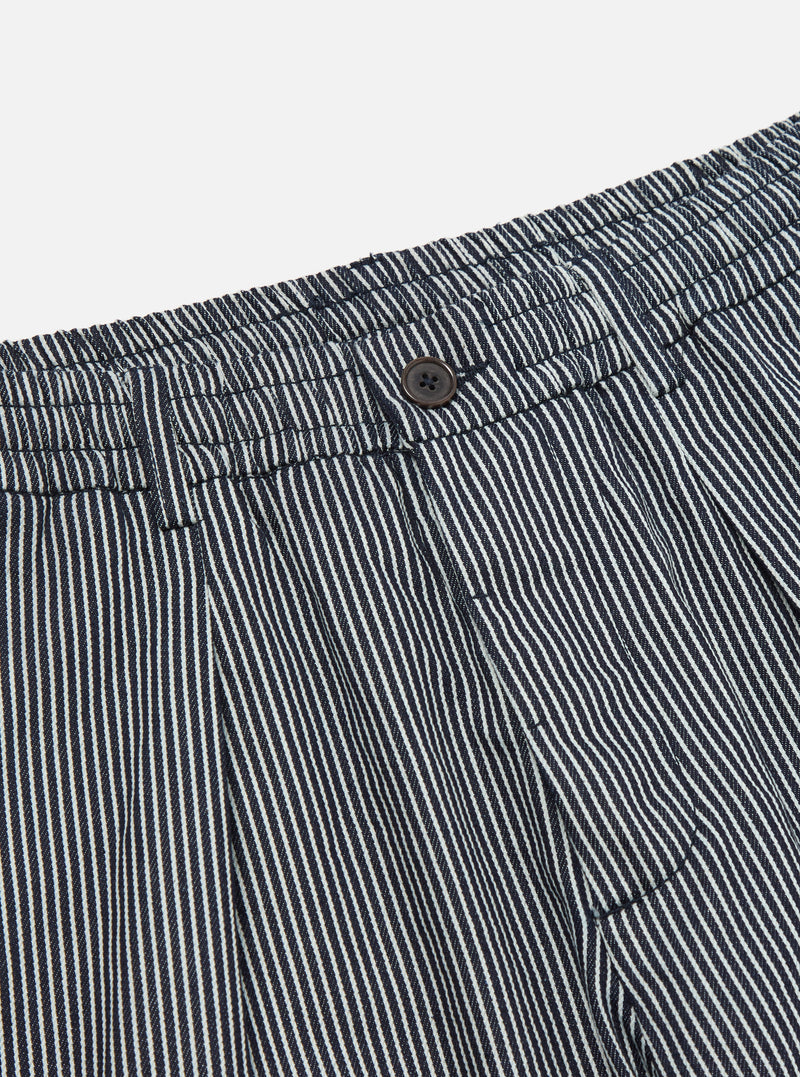 Close-up of the Indigo Hickory Stripe Denim Pleated Track Pant. Features include a button closure, pleated design, and denim material. Shot on a plain white background.