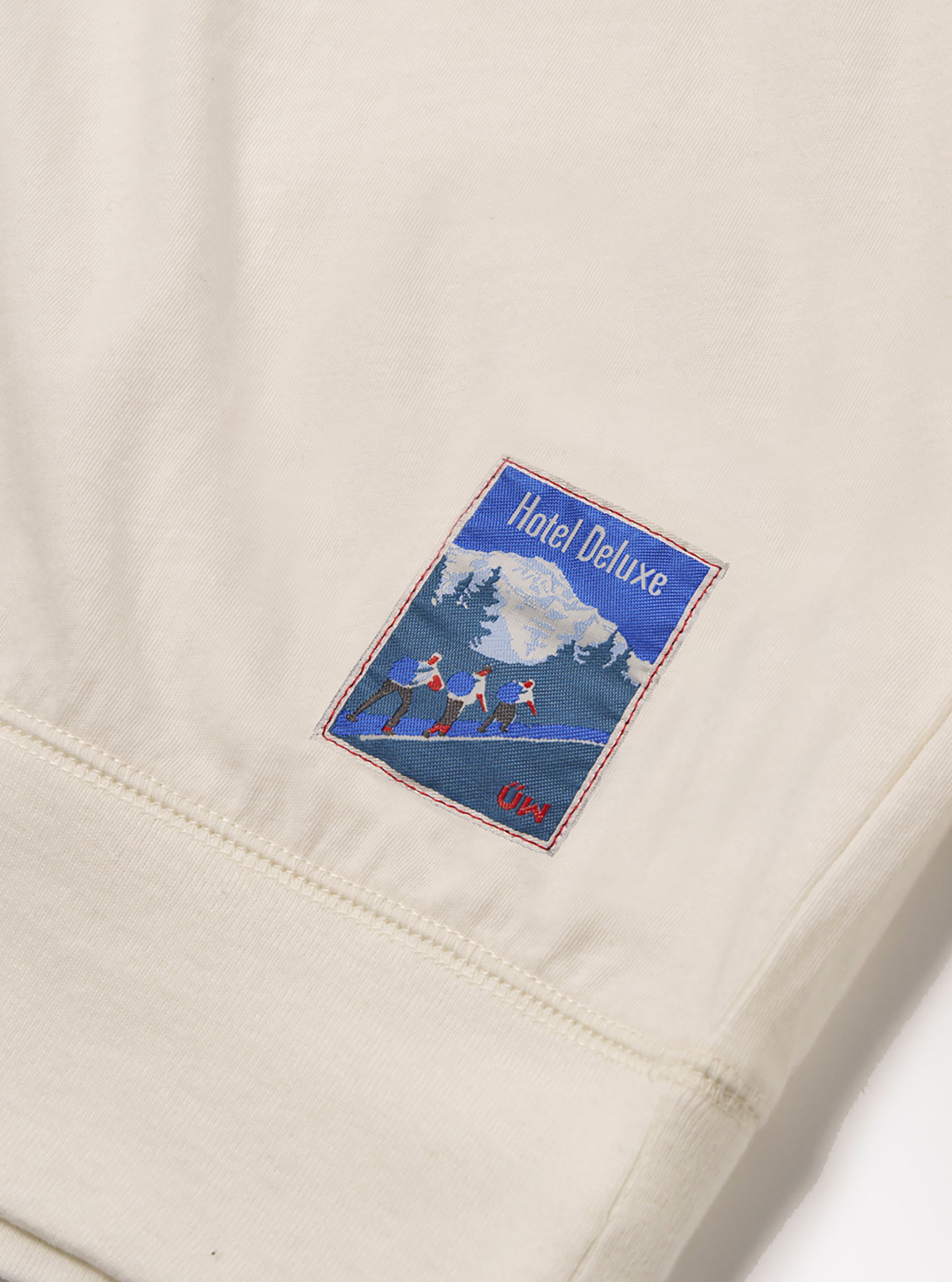Close up of the ecru Single Jersey Funnel Neck Tee showing the soft jersey material and a Hotel Deluxe patch. The tee is laid on a white surface.
