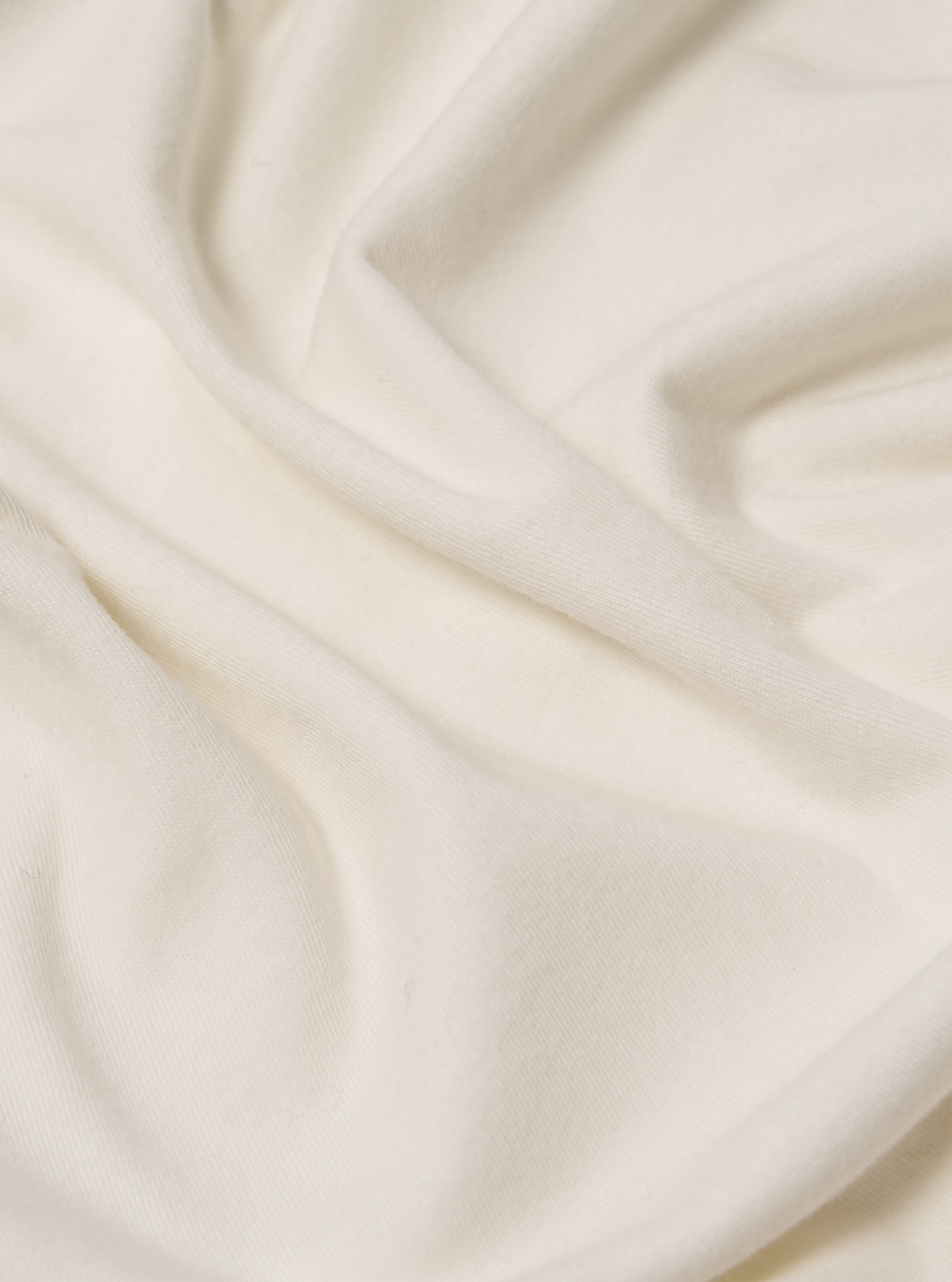 Close-up of the ecru Single Jersey Funnel Neck Tee fabric. The soft, single jersey material is draped on a plain surface, highlighting its texture and subtle ecru color.