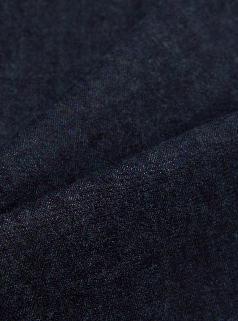 Close-up of the Indigo Brushed Denim Oxford Pant fabric. The indigo denim has a soft, brushed texture. Shot on a plain surface to highlight the material.