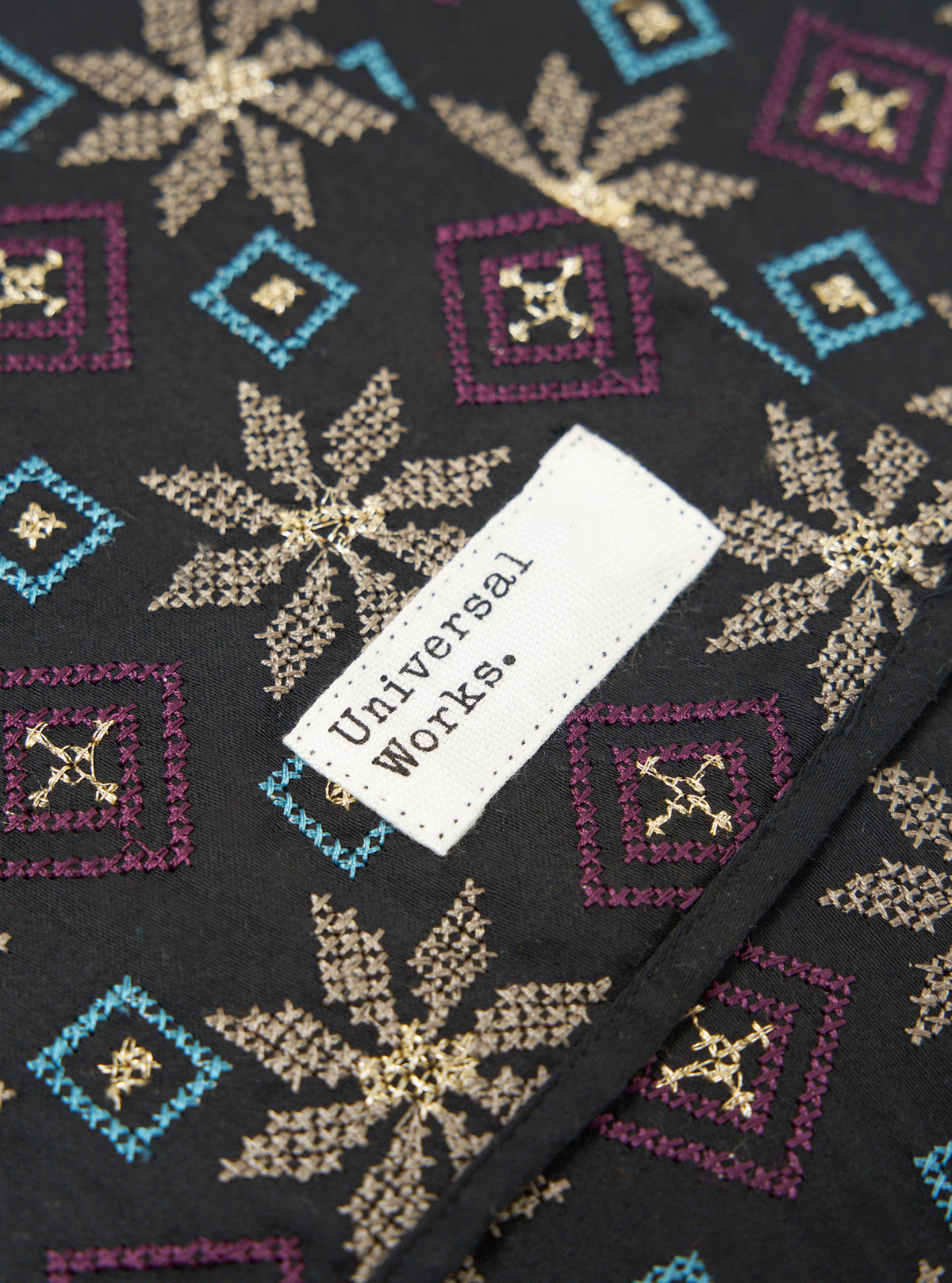 Close-up of the Black Embroidery Cotton Satin Embroidery Scarf. Features gold floral and blue and purple geometric embroidery on black cotton. A white tag is visible.