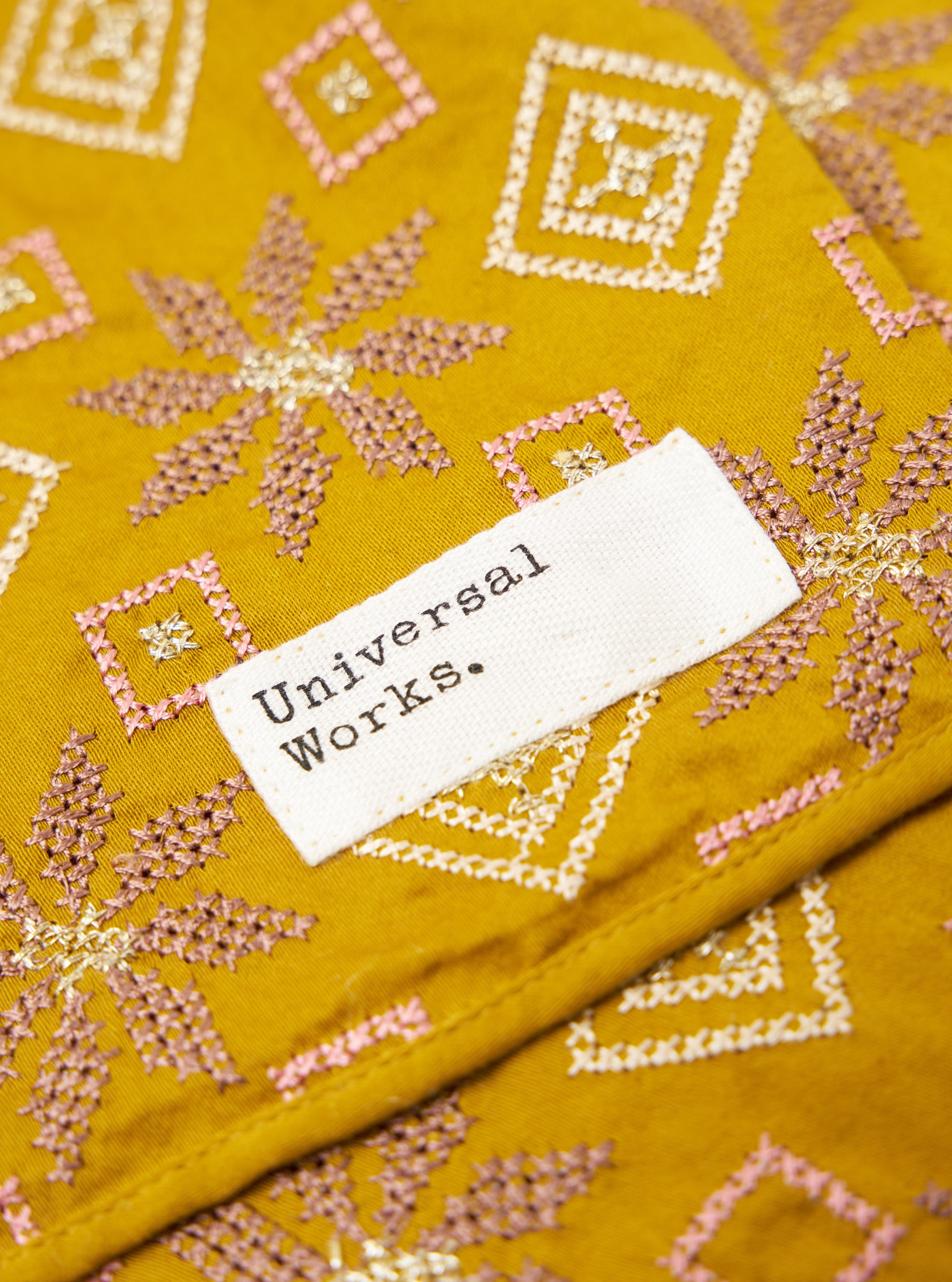 Close-up of the Mustard Embroidery Cotton Satin Embroidery Scarf. It features detailed pink, gold, and white embroidery on mustard fabric. A white tag with 'Universal Works.' is visible.