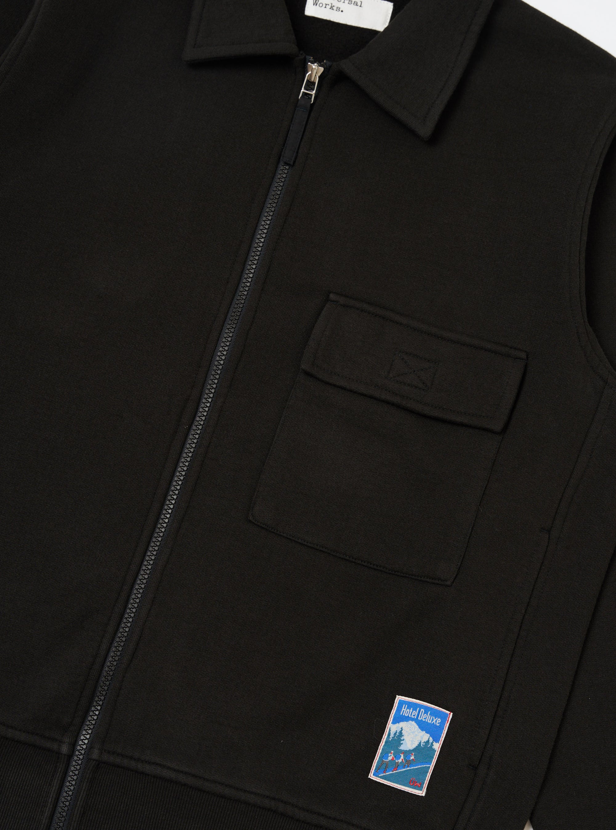 Close-up of the Black Brush Back Sweat Tyler Sweat Jacket. Features a collared neck, zip front, chest pocket, and Hotel Deluxe patch. Shot on a white background.