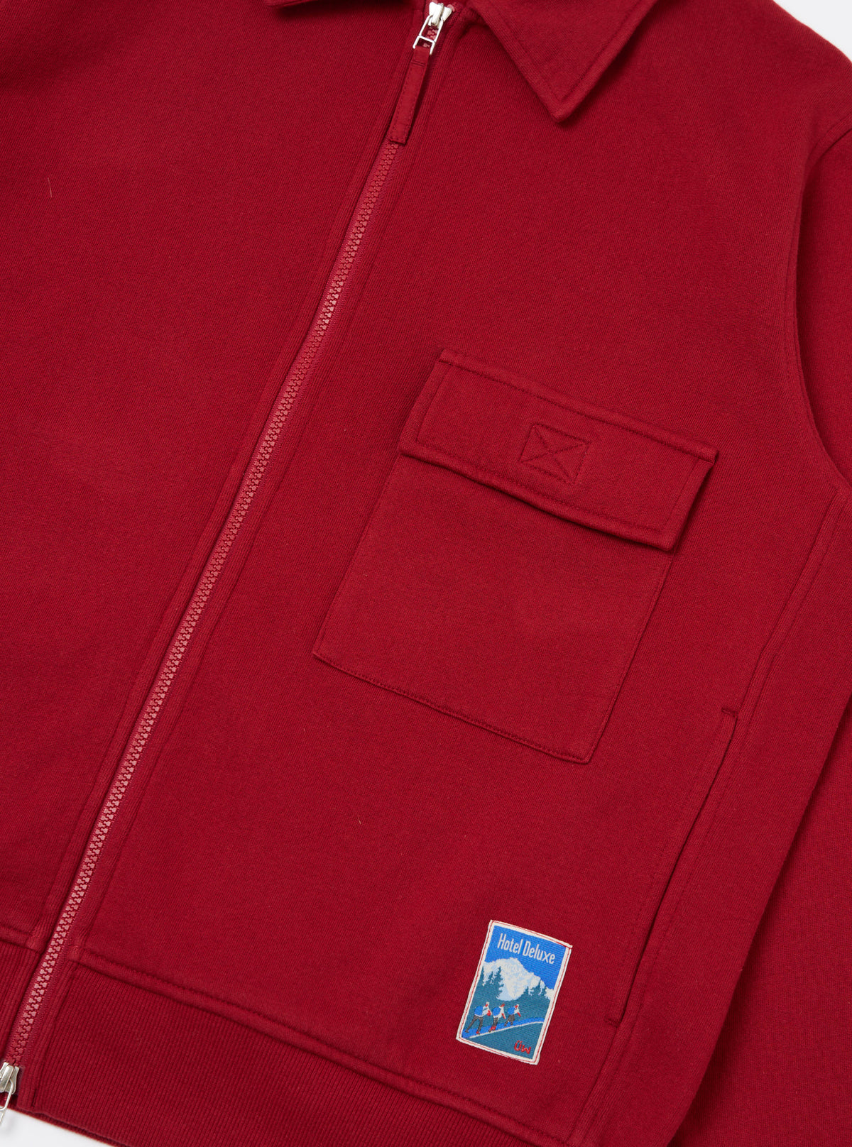 Close-up of the Red Brush Back Sweat Tyler Sweat Jacket. Features a zip front, chest pocket, and Hotel Deluxe patch. The jacket is displayed on a flat surface.