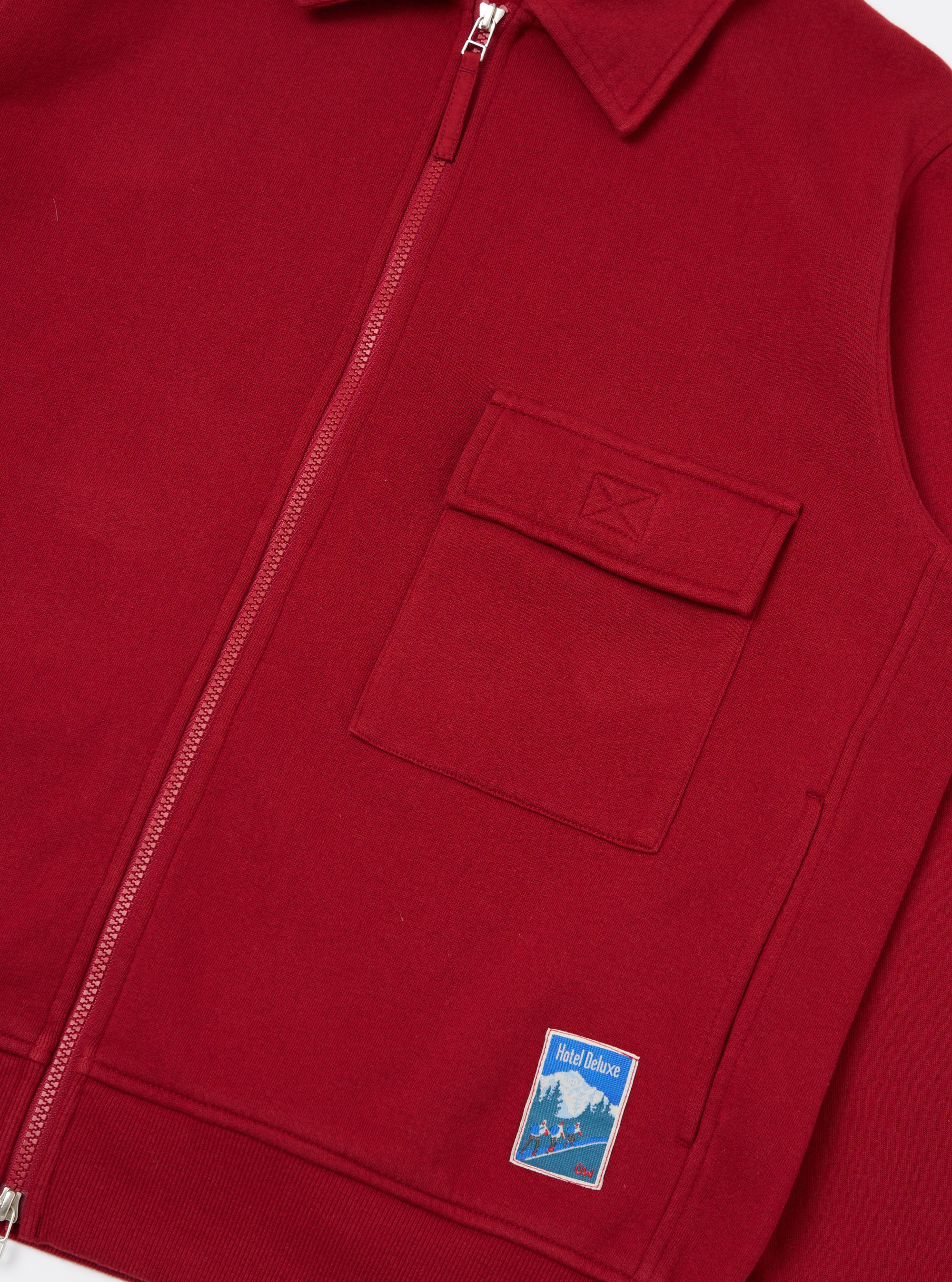 Close-up of the Red Brush Back Sweat Tyler Sweat Jacket. Features a zip front, chest pocket, and Hotel Deluxe patch. The jacket is displayed on a flat surface.
