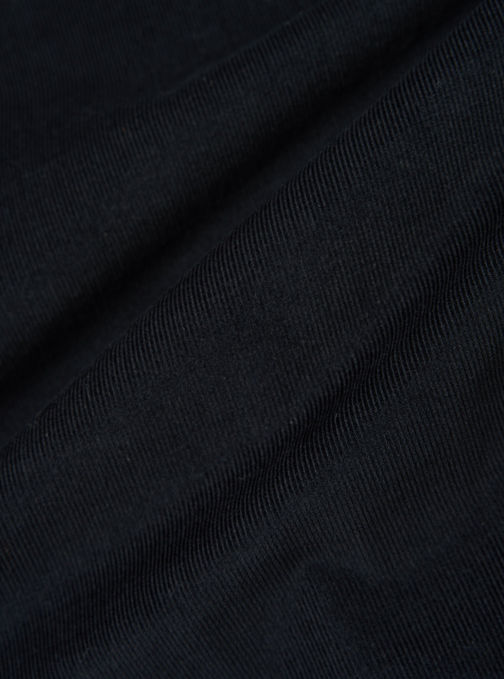 Close-up of the Black Organic Fine Cord Fatigue Shirt fabric. The fine cord texture is visible in the black material, draped on a flat surface.