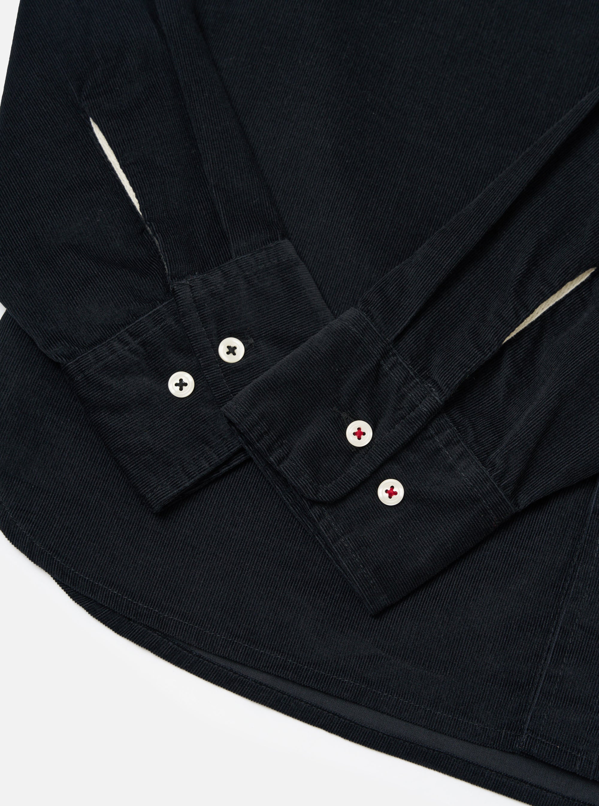 Close-up of the Black Organic Fine Cord Daybrook Shirt on a white surface. Shows the corduroy texture, buttoned cuffs, and curved hem.