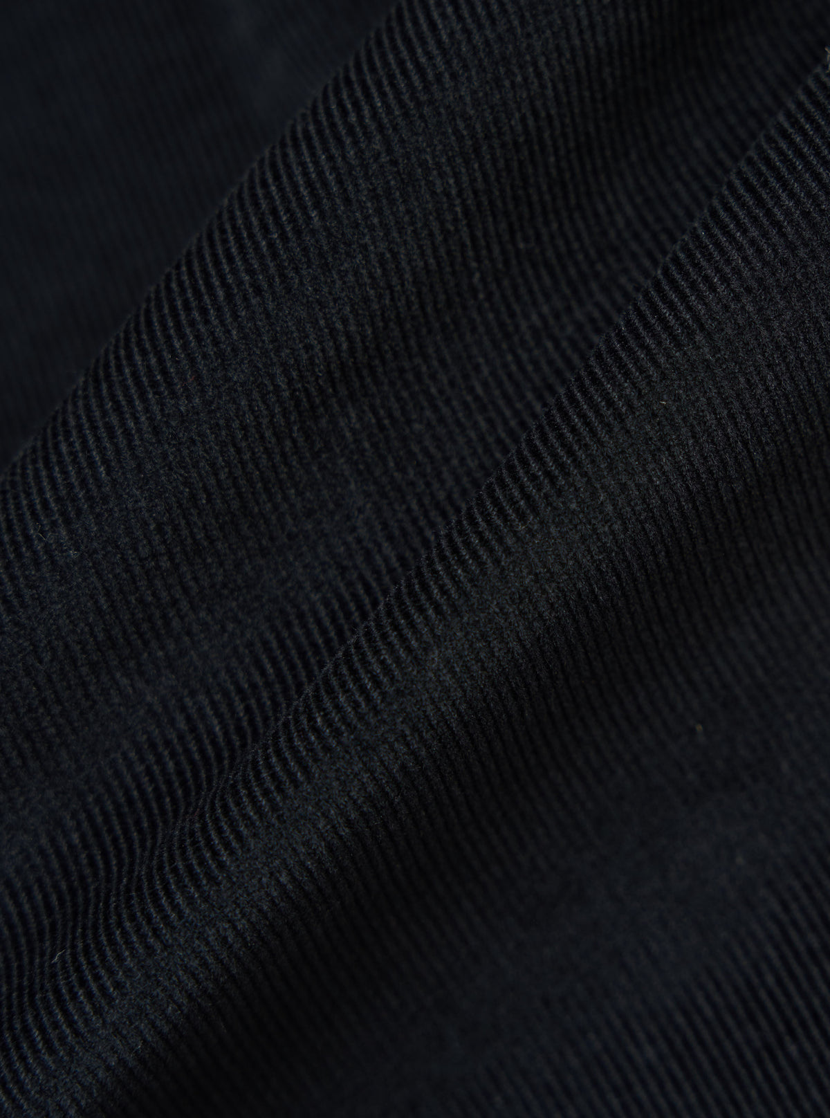 Close-up of the Black Organic Fine Cord Daybrook Shirt fabric. The dark, fine corduroy texture is softly lit, showing the material's depth and quality.