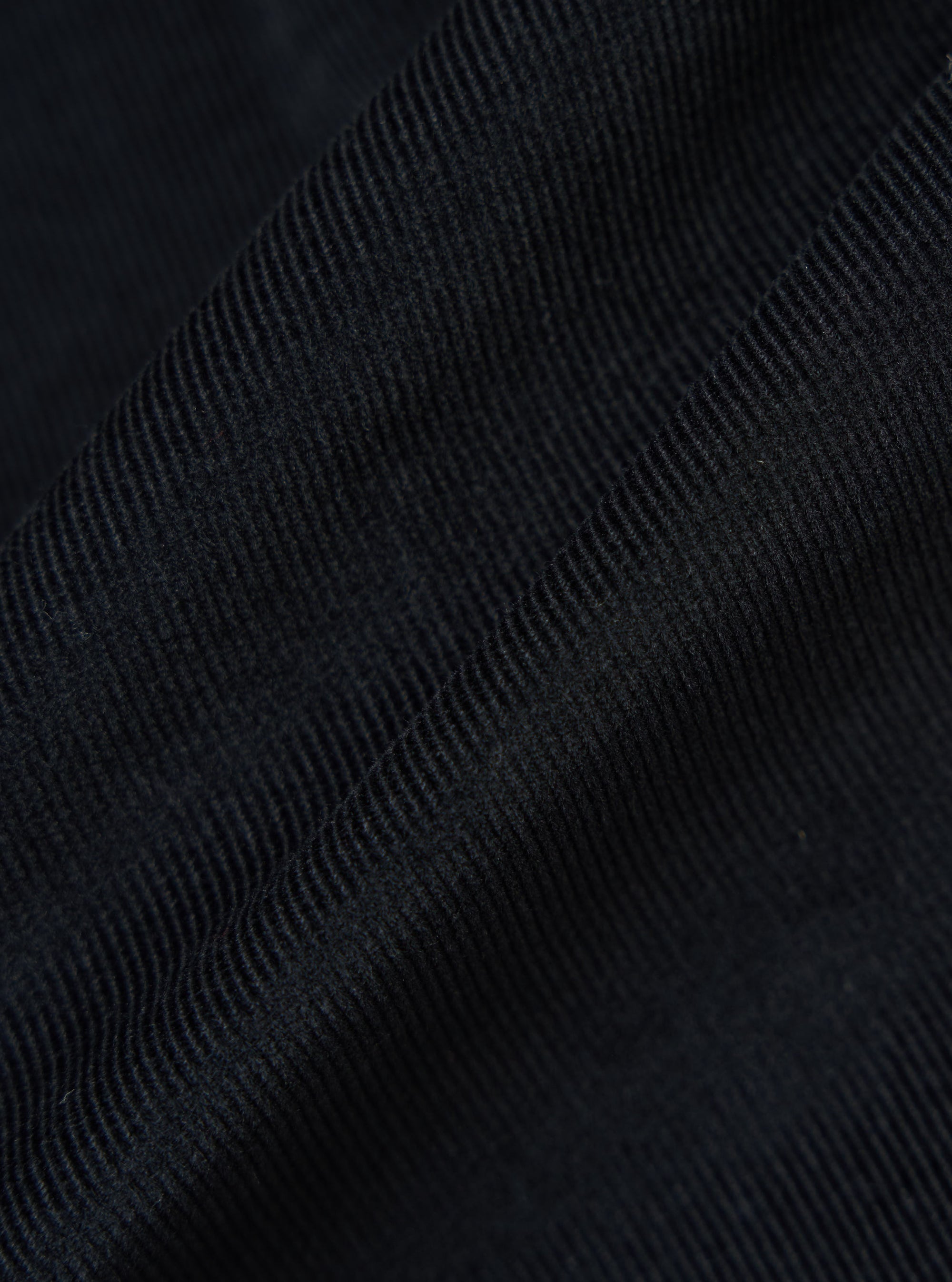 Close-up of the Black Organic Fine Cord Daybrook Shirt fabric. The dark, fine corduroy texture is softly lit, showing the material's depth and quality.