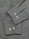 Close-up of the Charcoal Organic Chambray Patched Lazy Day Shirt, showing the charcoal chambray fabric, buttoned cuffs, and white buttons with red stitching. Shot on a flat surface.
