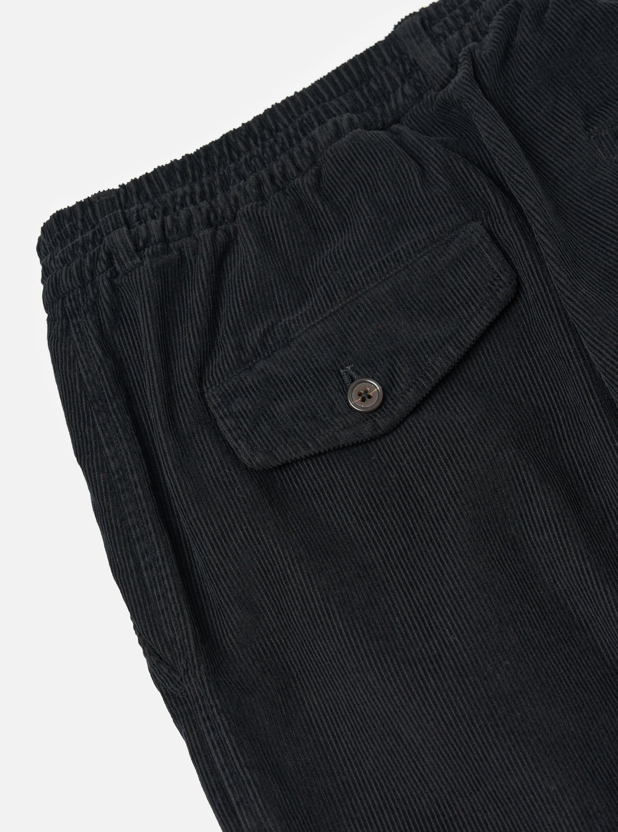 Close-up of the Black Cat Cord Pleated Track Pant, showcasing the black corduroy material, pleated design, elastic waistband, and back pocket with button closure on a white background.