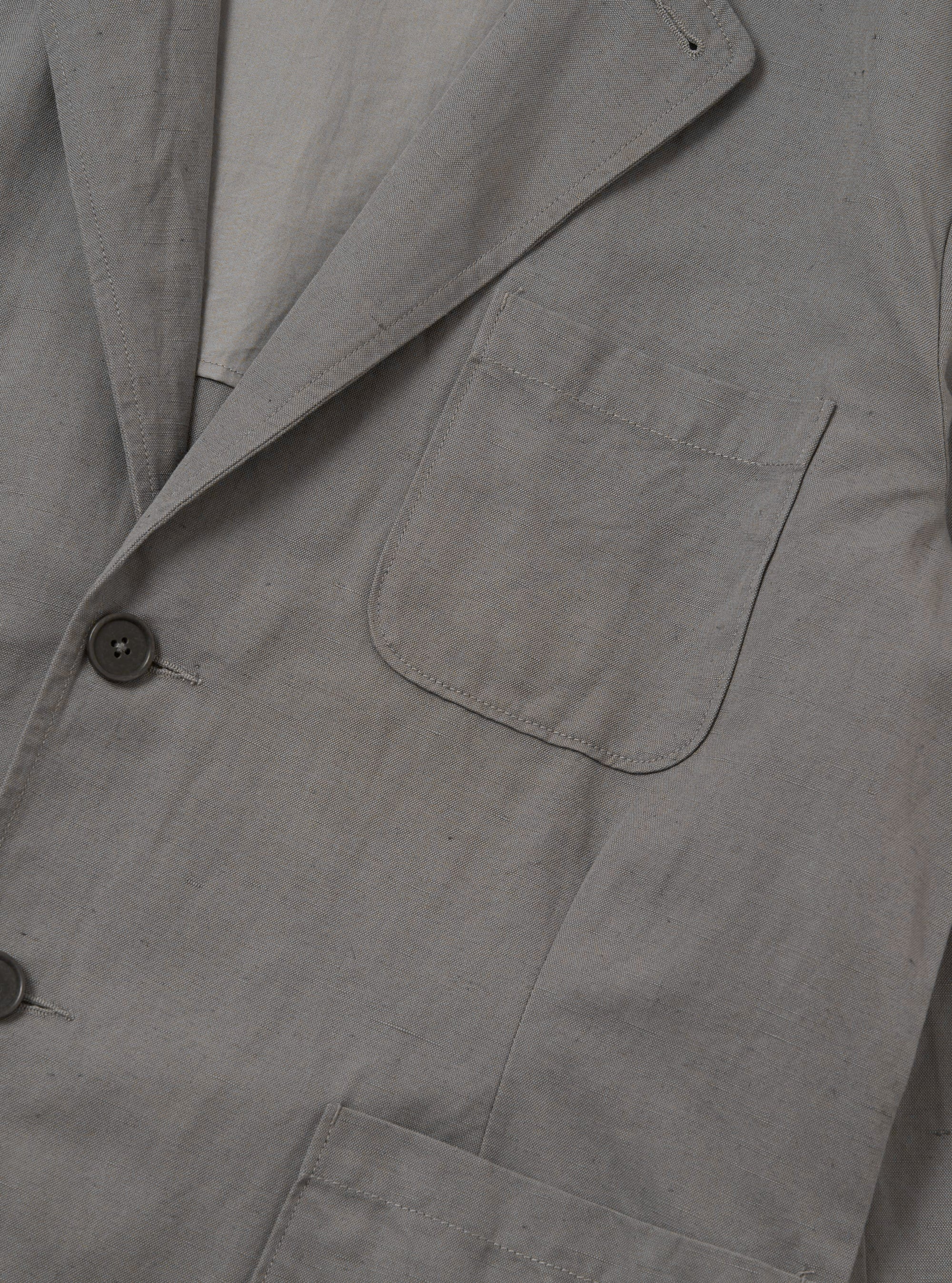 The Mid Grey Linen Slub Weave Two Button Jacket is shown as a flat lay against a white background. The jacket features a two-button front closure, a notched collar, and two large patch pockets at the hips. The fabric has a visible slub texture in a mid-grey tone.