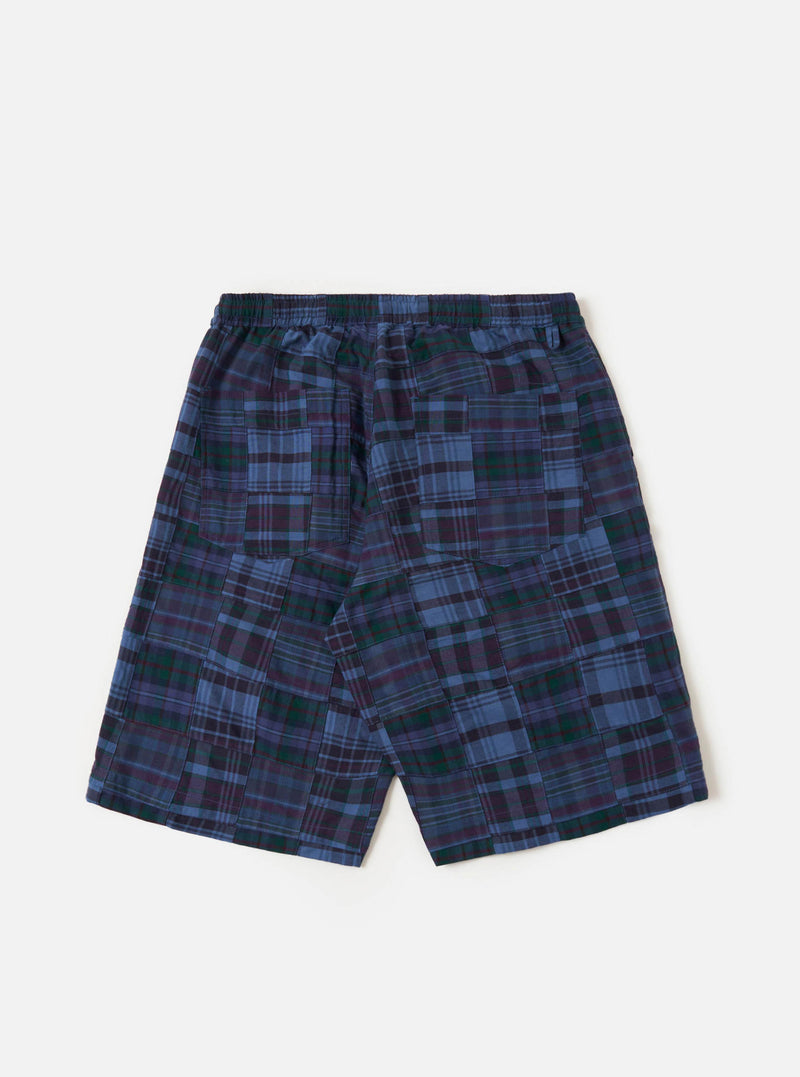 A pair of navy madras patchwork vacation shorts is shown from a rear angle, laid flat against a plain white background. The image highlights the patchwork construction of various blue and navy plaid patterns, the elasticated waistband with a drawstring, and a single rear patch pocket on the right side.