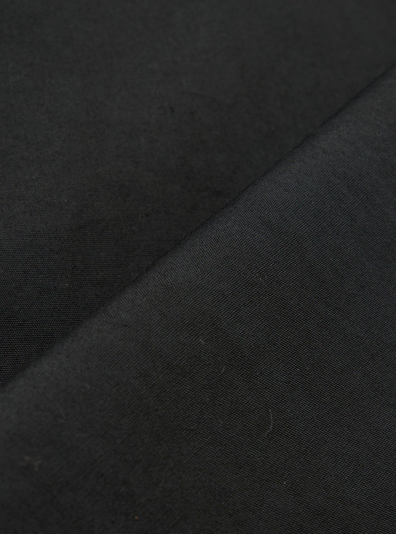 A close-up fabric swatch of the Black Broadcloth Oxford Pant shows a detailed view of the black broadcloth material. The image highlights the smooth, tightly woven texture of the cotton fabric against a neutral background.