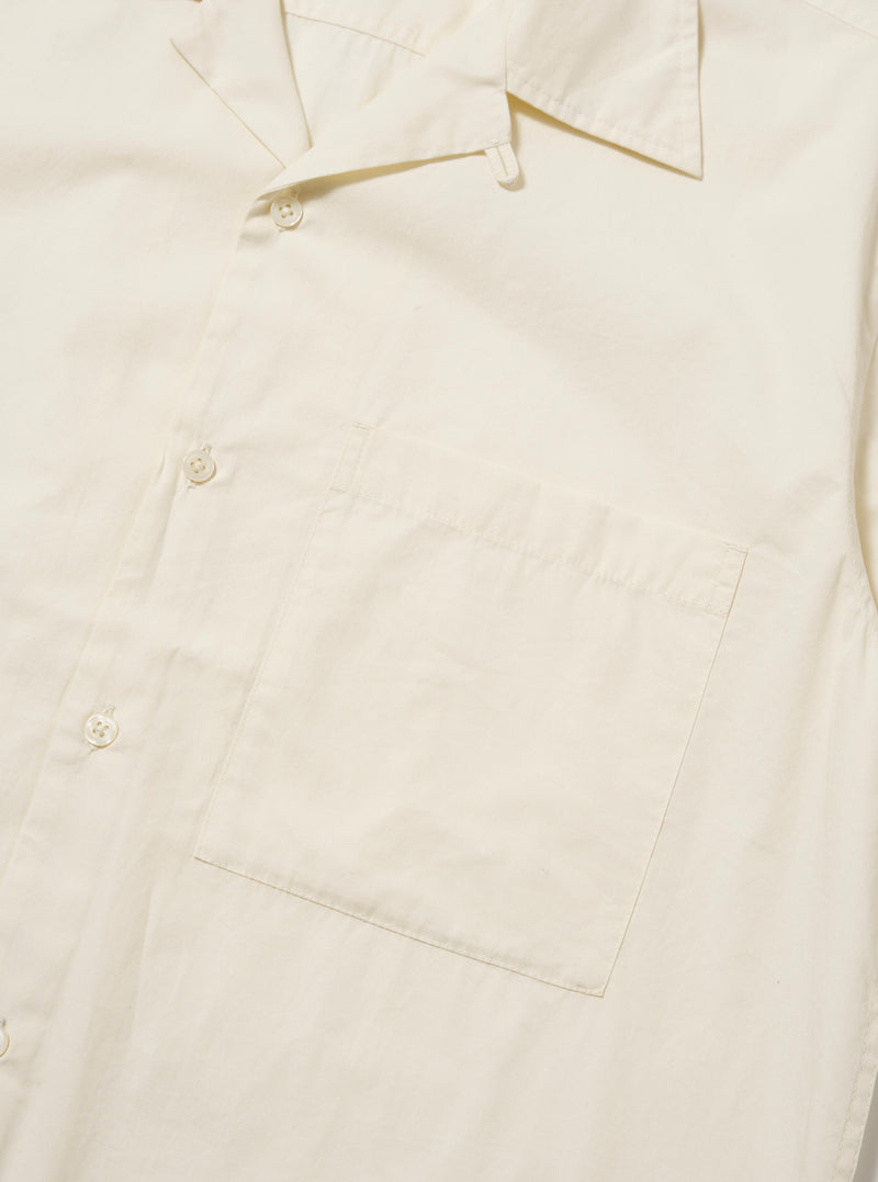 The Ecru Broadcloth Camp Shirt is shown from the back, laid flat against a plain white background. The image highlights the horizontal back yoke with two small pleats for added volume and the straight hem with side splits. The short sleeves are laid out to the sides, and