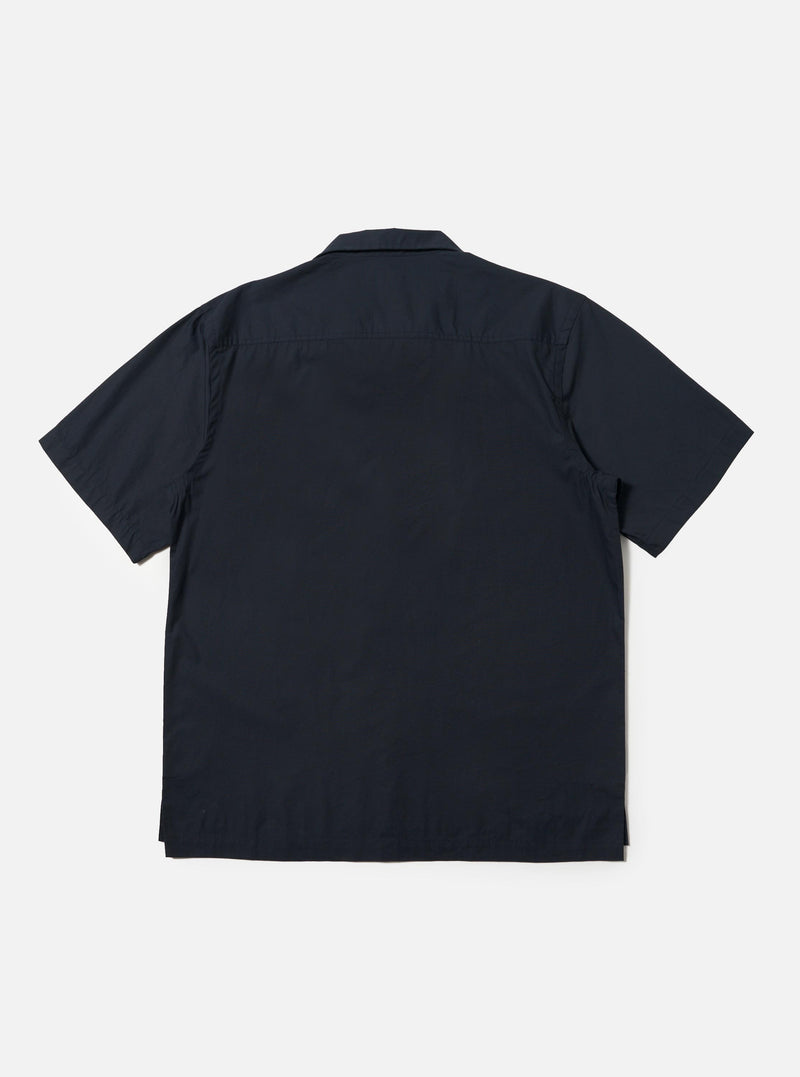 The Navy Broadcloth Camp Shirt is shown from the back, laid flat against a plain white background. The image highlights the back yoke with small pleats and the straight hem with side splits. The short-sleeved garment is made from a navy blue 100% cotton plain weave fabric.