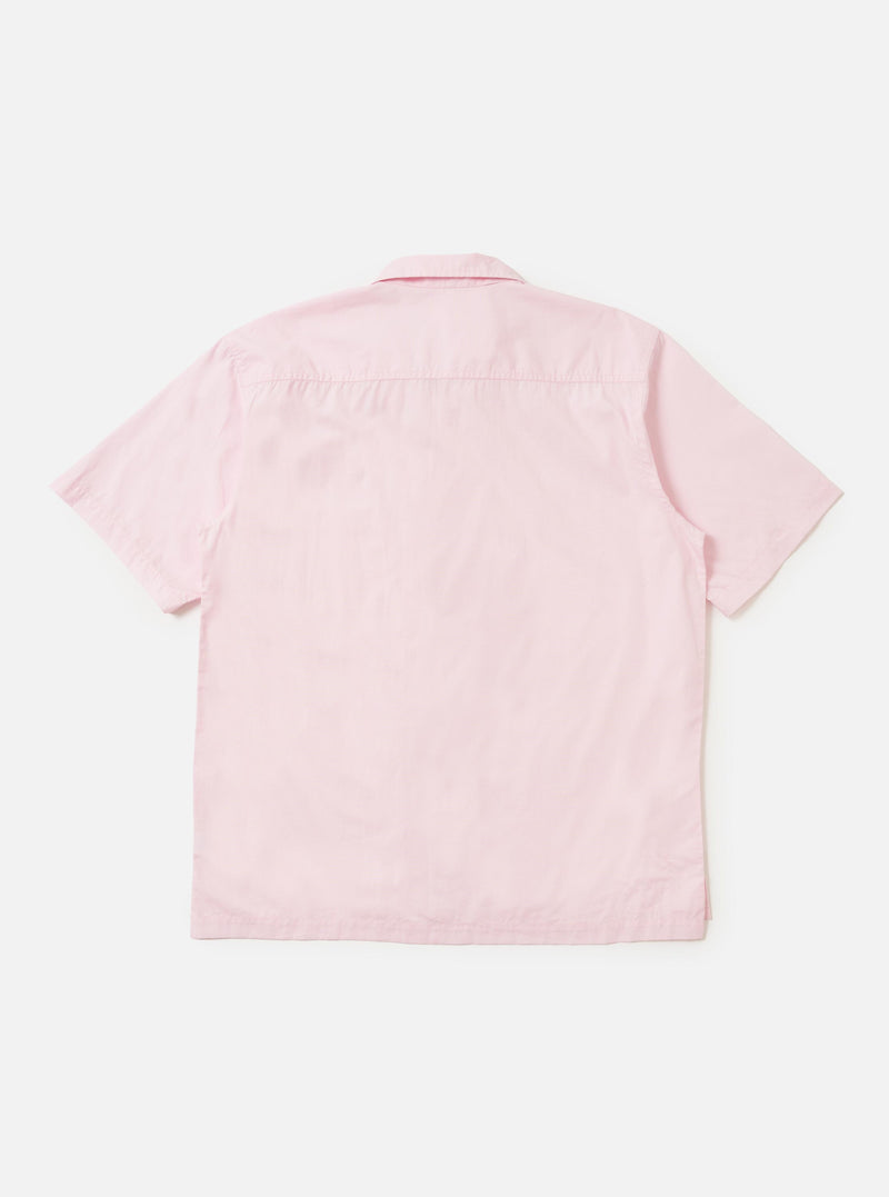 A model is wearing a Pale Pink Broadcloth Camp Shirt against a plain white background. The model is standing with one hand in their pocket, showcasing the loose, relaxed fit of the short-sleeved shirt. Prominent features include the open camp collar, a large chest pocket on the left side, and a straight hem. No mannequin is present in the image.