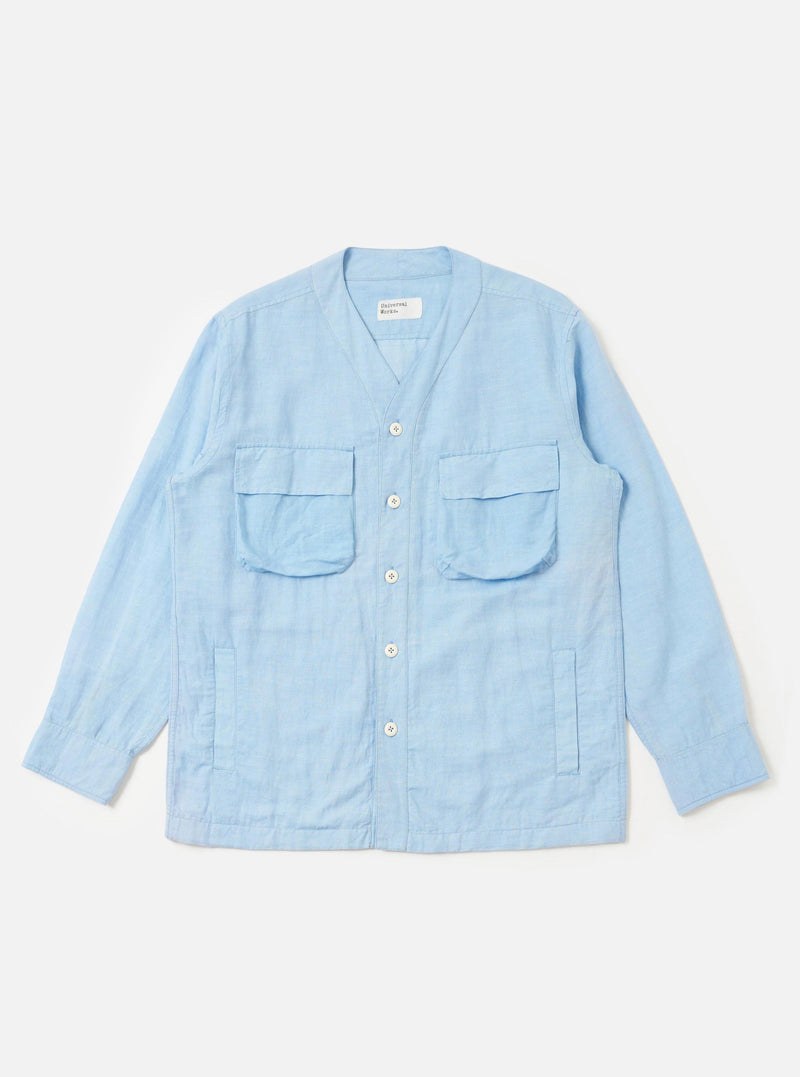 The Pale Blue Linen Cotton Shirting Parachute Shirt is displayed flat against a plain white background. This collarless, V-neck overshirt features a button-down front, two large 3D bellows pockets with buttoned flaps on the chest, and two vertical welt hand pockets near the waist. The garment is shown with long sleeves and a drawcord hem.