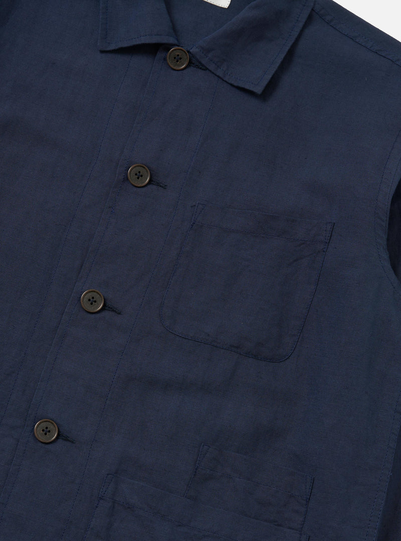 A model stands against a plain white background wearing the Navy Linen Cotton Shirting Bakers Overshirt over a white t-shirt. The navy overshirt features a classic collar, button-down front, and three large patch pockets on the front. The fabric has a visible linen texture, and the model has their hands by their sides.
