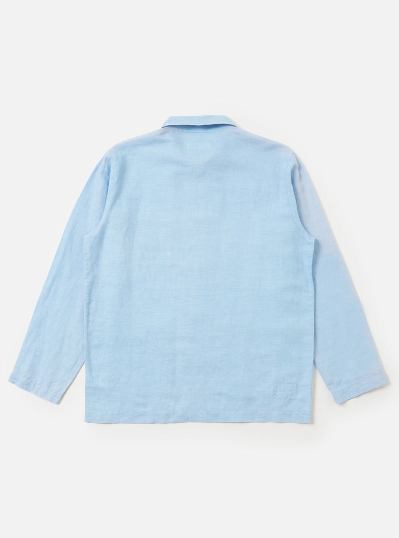 The Pale Blue Linen Cotton Shirting Bakers Overshirt is shown from the back, displayed flat against a plain white background. The image highlights the horizontal back yoke, simple one-piece collar, and the straight hem of the garment. The fabric is a light blue linen-cotton blend.