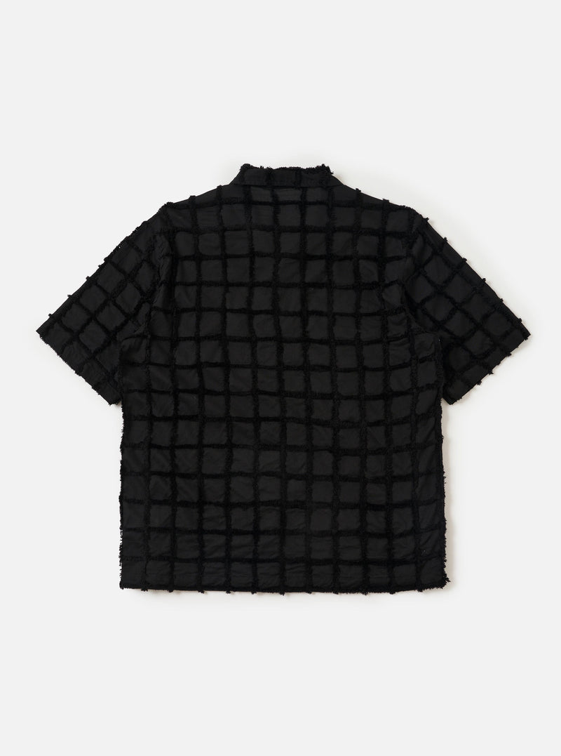 The Black 3D Check Road Shirt is shown from the back, laid flat against a solid white background. The image highlights the shirt's straight hem, short sleeves, and a horizontal yoke across the shoulders. The black fabric features a subtle, textured 3D check pattern throughout.