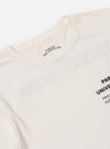 Close-up of the Paraboot x Universal Works Ecru Single Jersey Long Sleeve Tee on a white surface. The ecru tee features printed text. The label reads 'La Beaute Dans L'imperfection'.