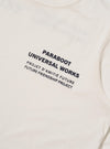Close-up of the Paraboot x Universal Works Ecru Single Jersey Long Sleeve Tee. The ecru tee features a printed logo. Shot on a flat surface.