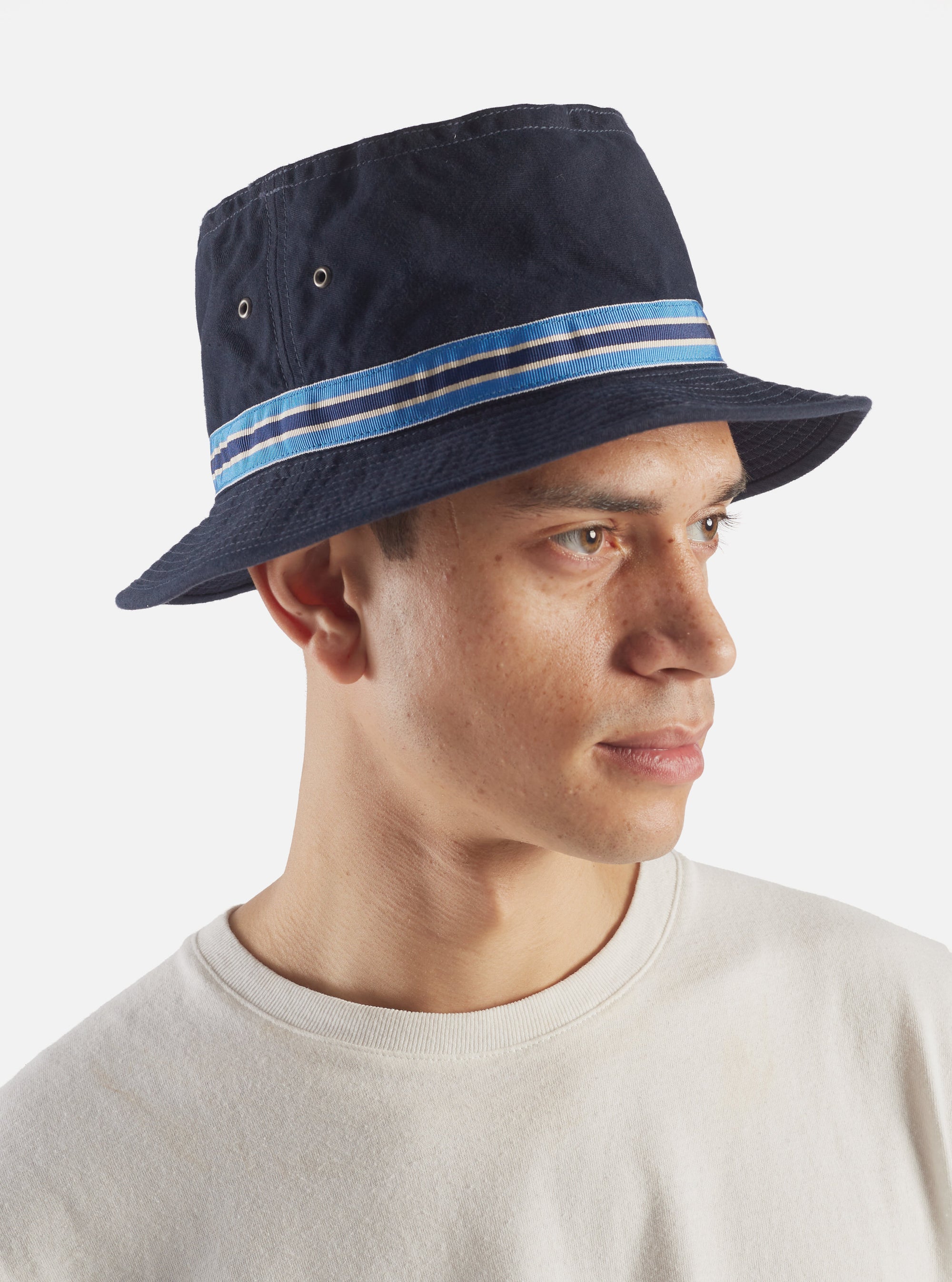 A model wears a cableami® Navy Chino Cotton Loose Light Pork Pie Hat. The hat is navy with a blue and white striped band. Shot against a white background.